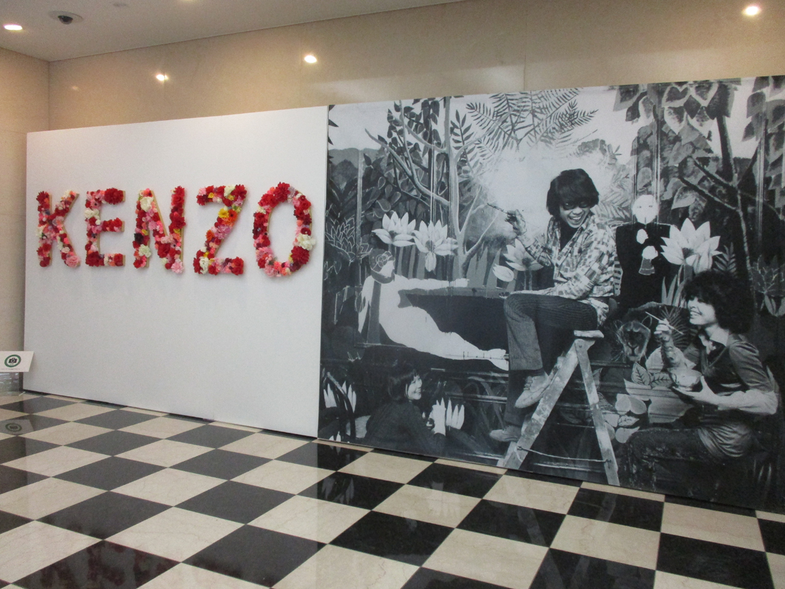 花で彩られたKENZOの文字と、若き日の高田賢三が最初の店の壁画を描いている巨大なモノクロ写真